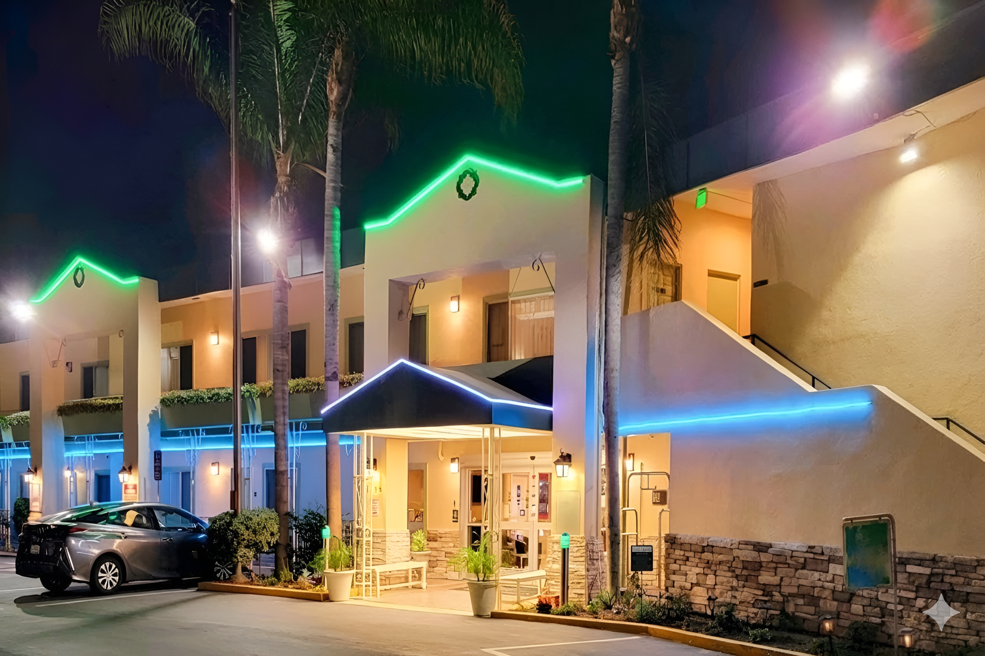 A motel with bright neon blue and green lights, palm trees, and a two-story building at night, inviting entrance under a blue awning.