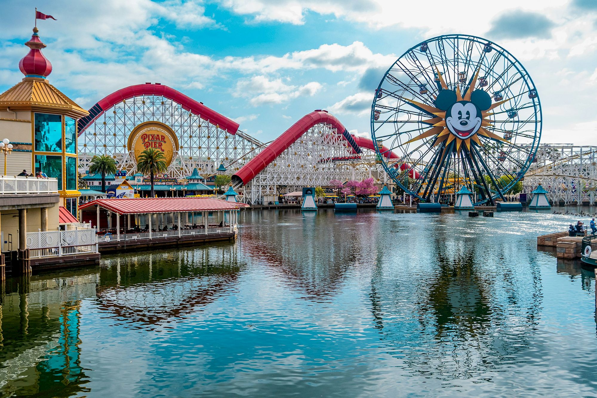 A lively seaside amusement pier with a pink roller coaster, arcade stalls, a large smiling Ferris wheel by the water, and reflections on a sunny harbor.