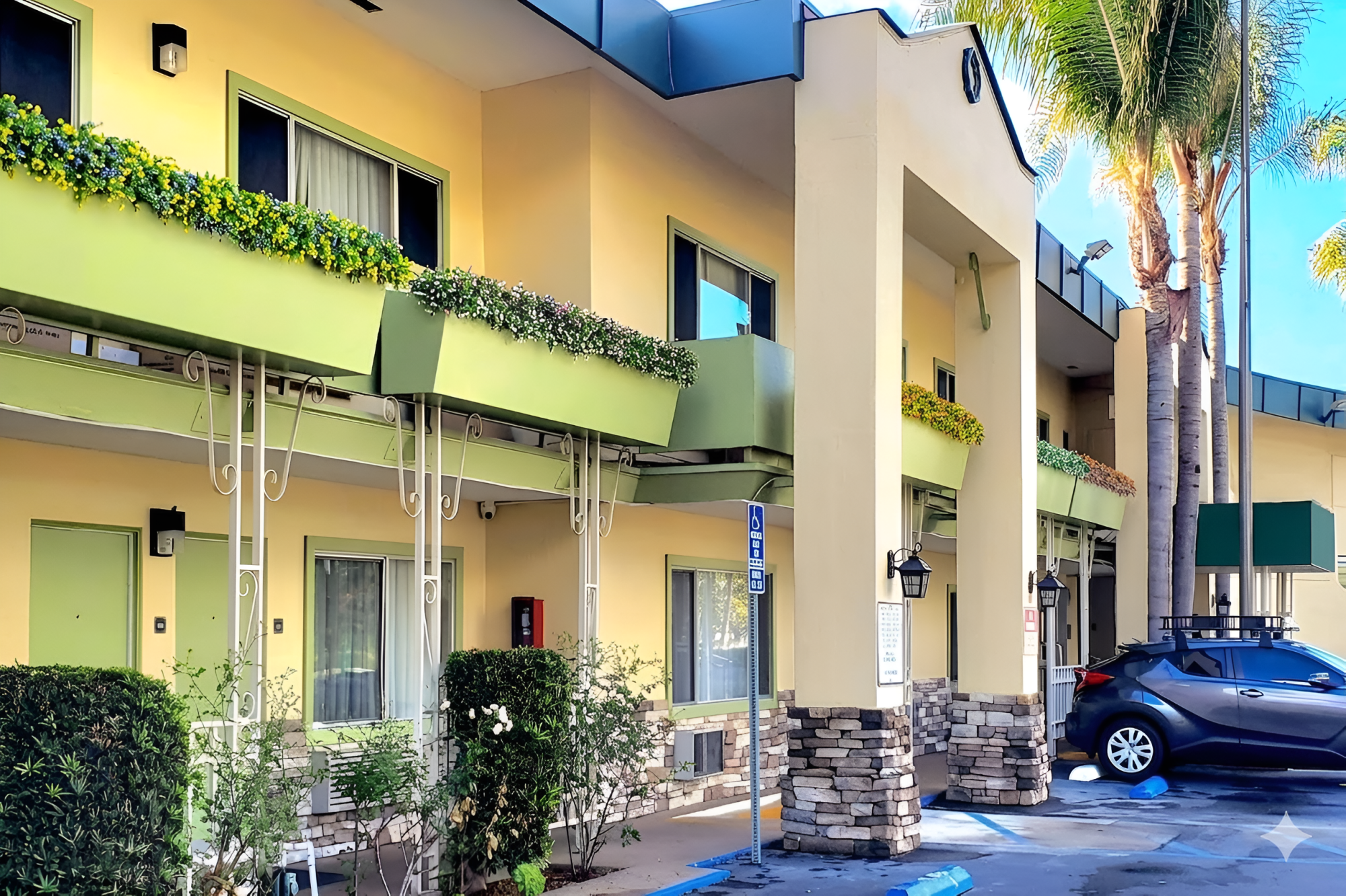 A colorful motel with green balconies, palm trees, a blue car, and a shaded walkway in a sunny, tropical setting.