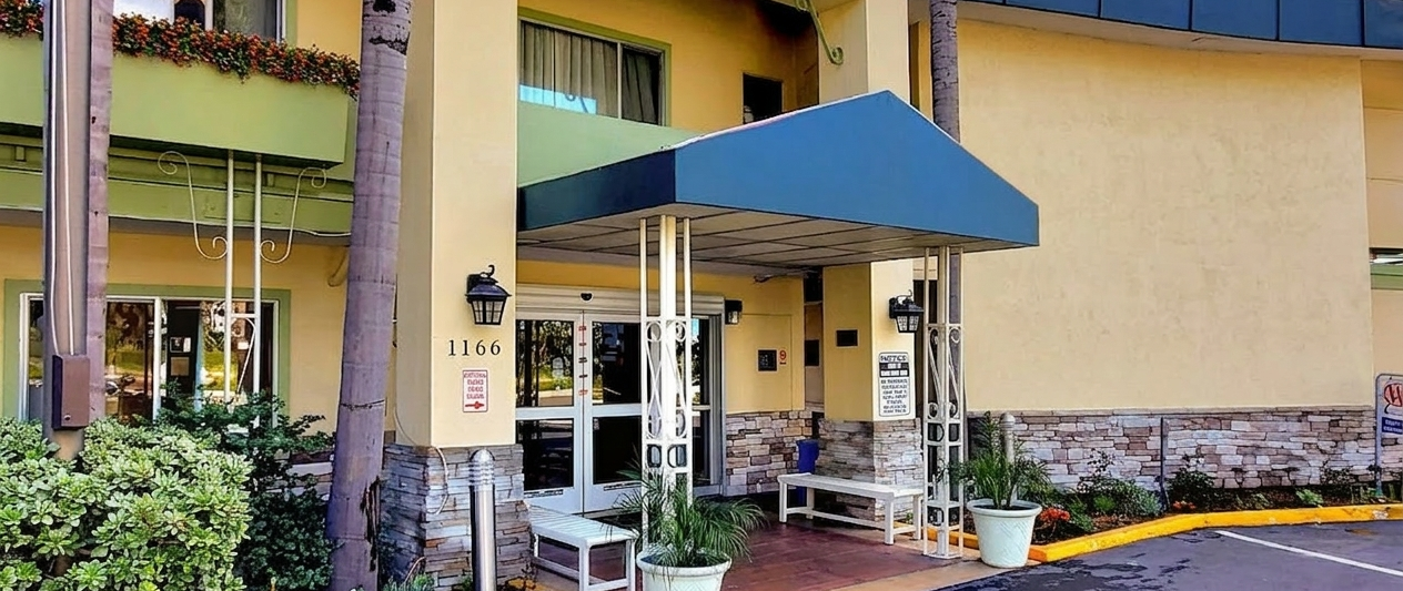 A motel entrance with a covered doorway, plants, and a small seating area under a blue awning by the parking lot.