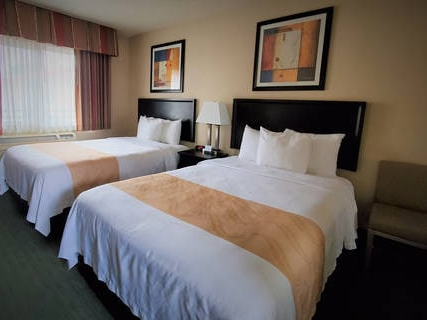Two double beds in a hotel room with beige bed runners, a nightstand, lamp, framed art, and a window with curtains ending the sentence.