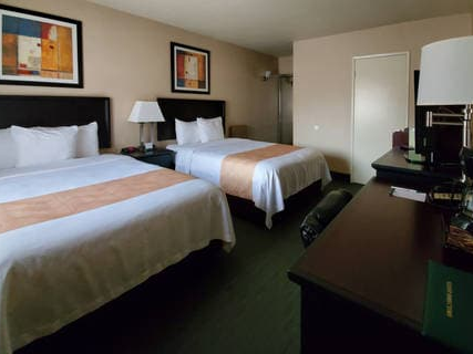 Two double beds with white linens and beige runners, a desk with a lamp, and wall art in a hotel room.