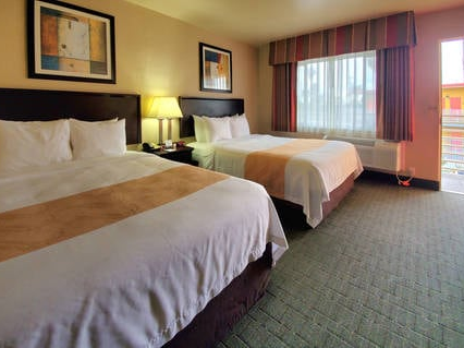 Two beds in a hotel room with lamps between, a window with red curtains, and an outside view; cozy and simple.