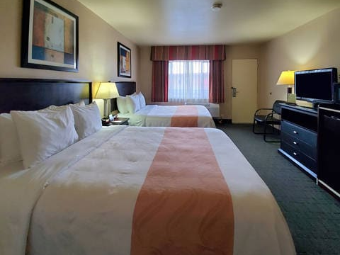 Two double beds with white linens and peach runners, a desk with a TV, lamps, a window with red curtains, and framed art on the wall.