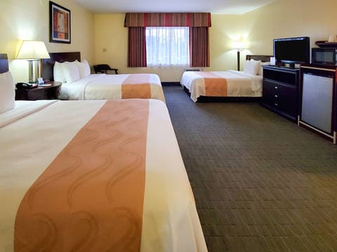 A hotel room with three beds, warm lighting, a window with drapes, a desk and TV, and a muted orange-brown runner on each bed.