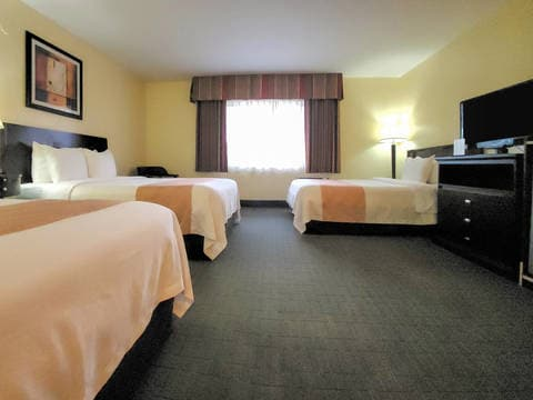 A hotel room with three beds, a dresser, TV, and a window with curtains; soft lighting and neutral decor, spacious and tidy.