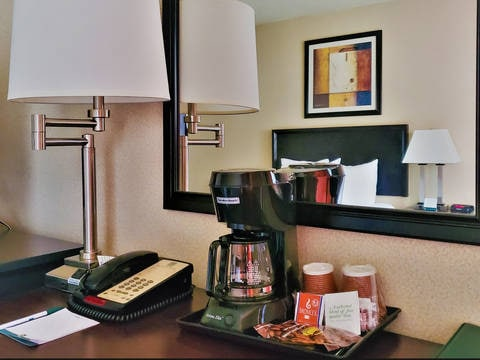 A hotel desk with a coffee maker, two lamps, a phone, a tray of condiments, and a mirror reflecting a framed artwork above the bed.