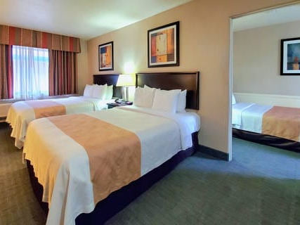 Hotel room with two large beds, warm lighting, framed art, and a connected smaller room visible through an open doorway.