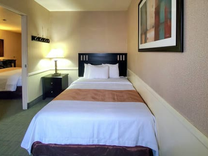 A tidy hotel room with a single bed, nightstand, lamp, framed art, and a view into an adjacent room with a mirror and another bed, neatly arranged.