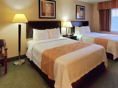 Two queens hotel room with two beds, lamps, nightstands, and a window view, neatly made with beige bed runners.