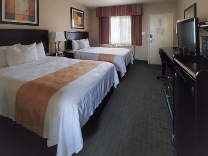 Three beds in a hotel room with nightstands, lamps, a window, a desk, and a television across from the beds, neutral decor.