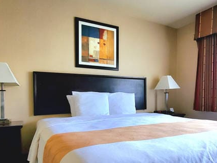 A neatly made hotel bed with white linens and a tan runner, two nightstands with lamps, a framed abstract art piece above the headboard, beige walls, and a window with red curtains.
