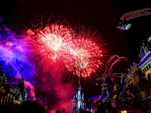 Fireworks explode over a festive street at night, with colorful lights, smoke, and cheering crowds beneath illuminated buildings.