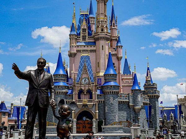 Disney's Cinderella Castle at Magic Kingdom, with a statue of Walt and Mickey in the foreground, bright flowers, and visitors nearby.
