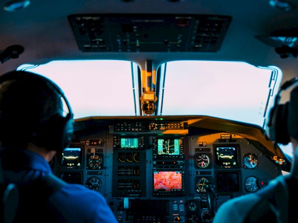 Two pilots in a cockpit with screens and controls, preparing for flight, seen from behind, with an illuminated cockpit dashboard in view.
