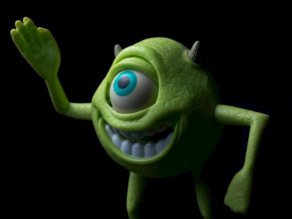 A green one-eyed monster from Monsters, Inc., waving hello with a goofy grin on a black background.