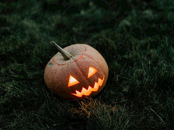 A glowing carved pumpkin with a jagged grin sits on dark grass, lit from inside, casting a spooky orange glow.
