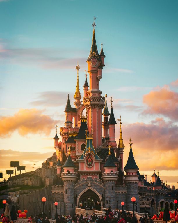 A fairy-tale castle with tall spires rises against a sunset sky, crowds gathered at the base, lanterns glowing as day fades.