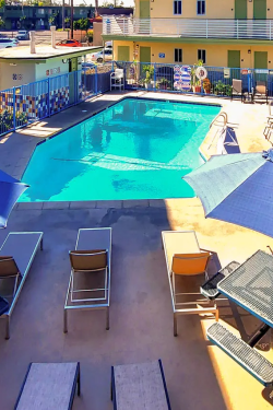 A rectangular hotel pool with blue water, surrounded by lounge chairs, umbrellas, and a sunny courtyard in a pastel-colored apartment complex.