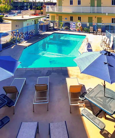 A rectangular hotel pool with blue water, surrounded by lounge chairs, umbrellas, and a sunny courtyard in a pastel-colored apartment complex.