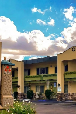 A row of beige motel-style buildings with green balconies, a curved wall in the foreground, blue sky with white clouds, and a parked car.
