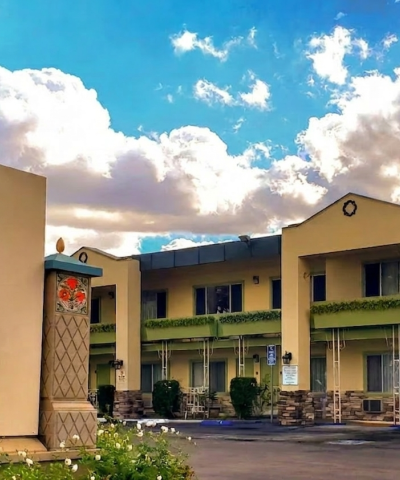 A row of beige motel-style buildings with green balconies, a curved wall in the foreground, blue sky with white clouds, and a parked car.