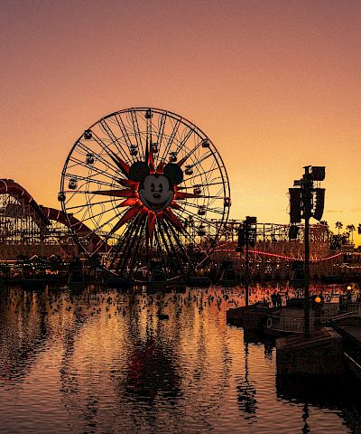 A twilight view of an amusement pier with a glowing ferris wheel, roller coaster, and water reflections along the harbor, in Orange and purple skies.