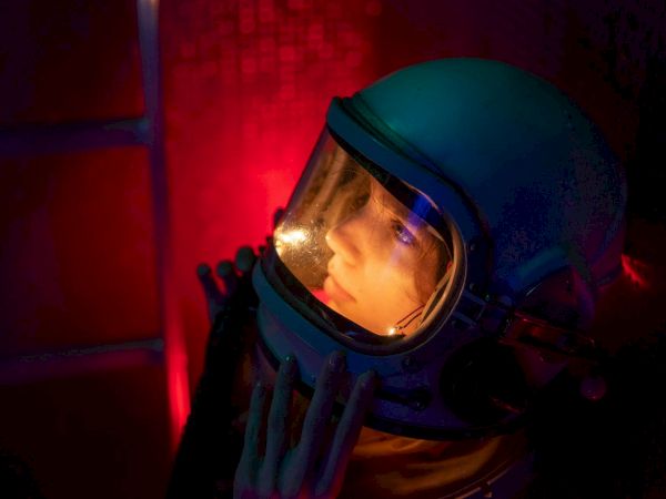 A person wearing a blue helmet with a clear visor, illuminated by red light, looking upward with a gloved hand near the face.