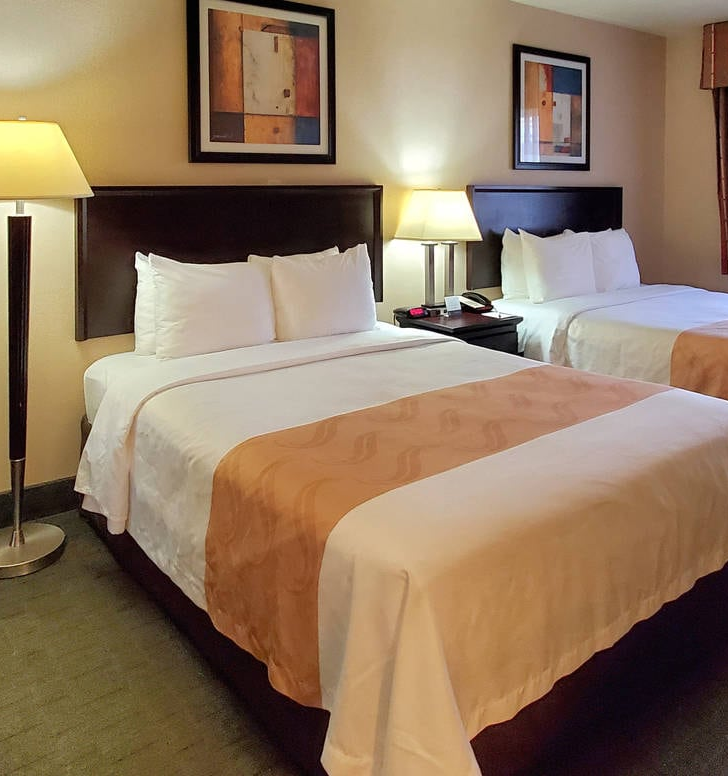 A hotel room with two double beds, beige runners, two lamps, a chair, framed art, a desk with a phone, and a window with blinds.