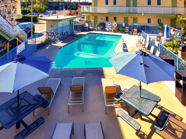 Poolside at a sunny resort with blue umbrellas, lounge chairs, a rectangular pool, and a pastel yellow building in the background.
