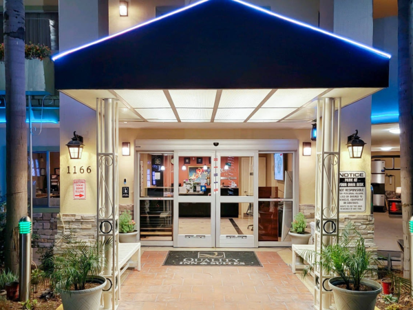 People entering a hotel or lobby beneath a blue-lit awning; glass doors, potted plants, and warm interior lighting at the entrance.