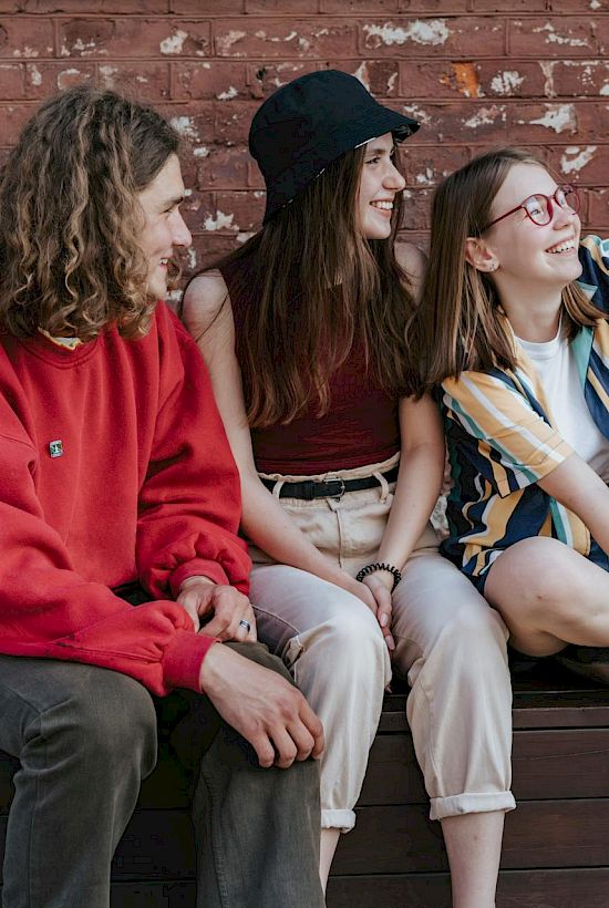 Three friends sit on a bench by a brick wall, taking a selfie together.