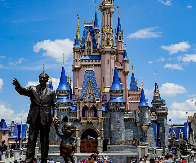A grand castle with blue spires stands behind a statue and a crowd in front, under a bright blue sky with fluffy clouds.