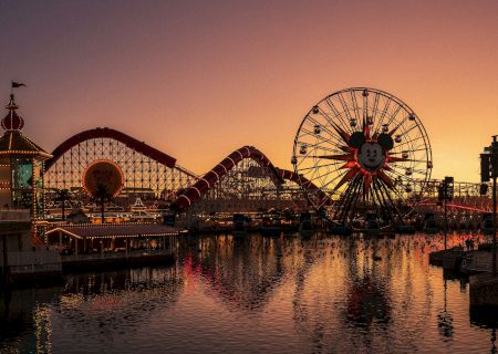A sunset over an amusement pier with a roller coaster and a lit Ferris wheel reflecting on calm water.