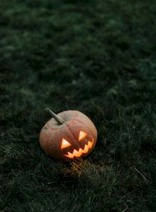 A glowing carved pumpkin with a jagged grin sits on dark grass, lit from inside, creating a spooky Halloween vibe.