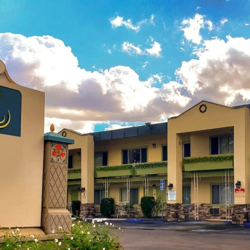 Colorful roadside motel with yellow/beige buildings, a curved sign, green hedges, and a blue sky with fluffy clouds, parking lot, and a car.