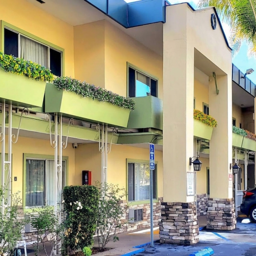A bright, two-story yellow building with balconies, palm trees, and parked scooters in a sunny courtyard.
