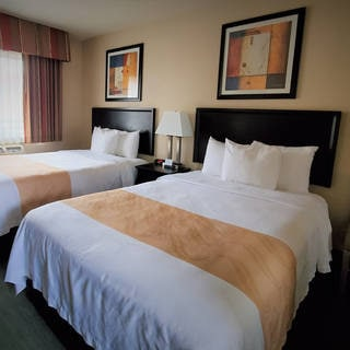 Two double beds with white linens and tan runners, a nightstand with lamp between them, framed art, a window with curtains, and a beige chair in a hotel room.