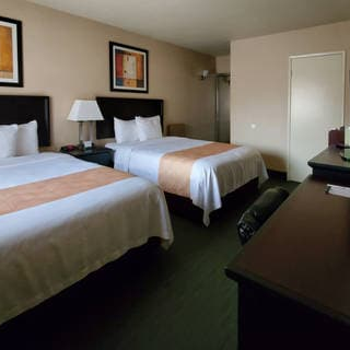 Two queen beds in a hotel room, with matching lamps, framed art, a desk and chair, and a doorway at the back; neutral beige walls.