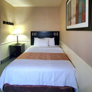 A small hotel room with a single bed, dark headboard, bedside lamp, framed artwork, and a doorway to an adjacent room; simple decor.