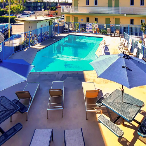 A sunny hotel pool area with blue umbrellas, lounge chairs, a rectangular pool, and surrounding buildings; people relaxing by the water.