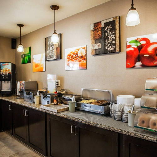 A caf&eacute; or buffet smoothie bar setup with coffee machines, plates, and fruit-themed wall art, clean counters, warm lighting, and storage cabinets.