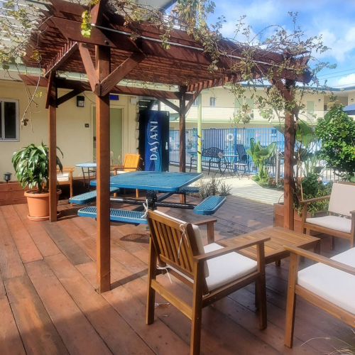 A wooden deck with seating, a pergola draped in vines, outdoor chairs and a table, potted plants, and a blue umbrella in a sunny courtyard.