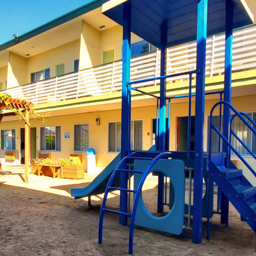 A sunny playground outside a two-story motel/complex with blue metal stairs, slide, and a picnic area under a pergola, sand ground, and balconies.