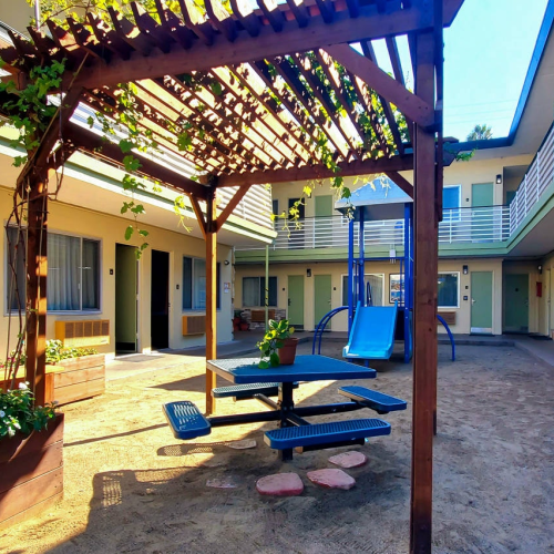 A sunny courtyard of a motel or dorm with a wooden pergola, picnic table, blue slide, potted plants, and a few benches along beige stucco walls, outdoors.