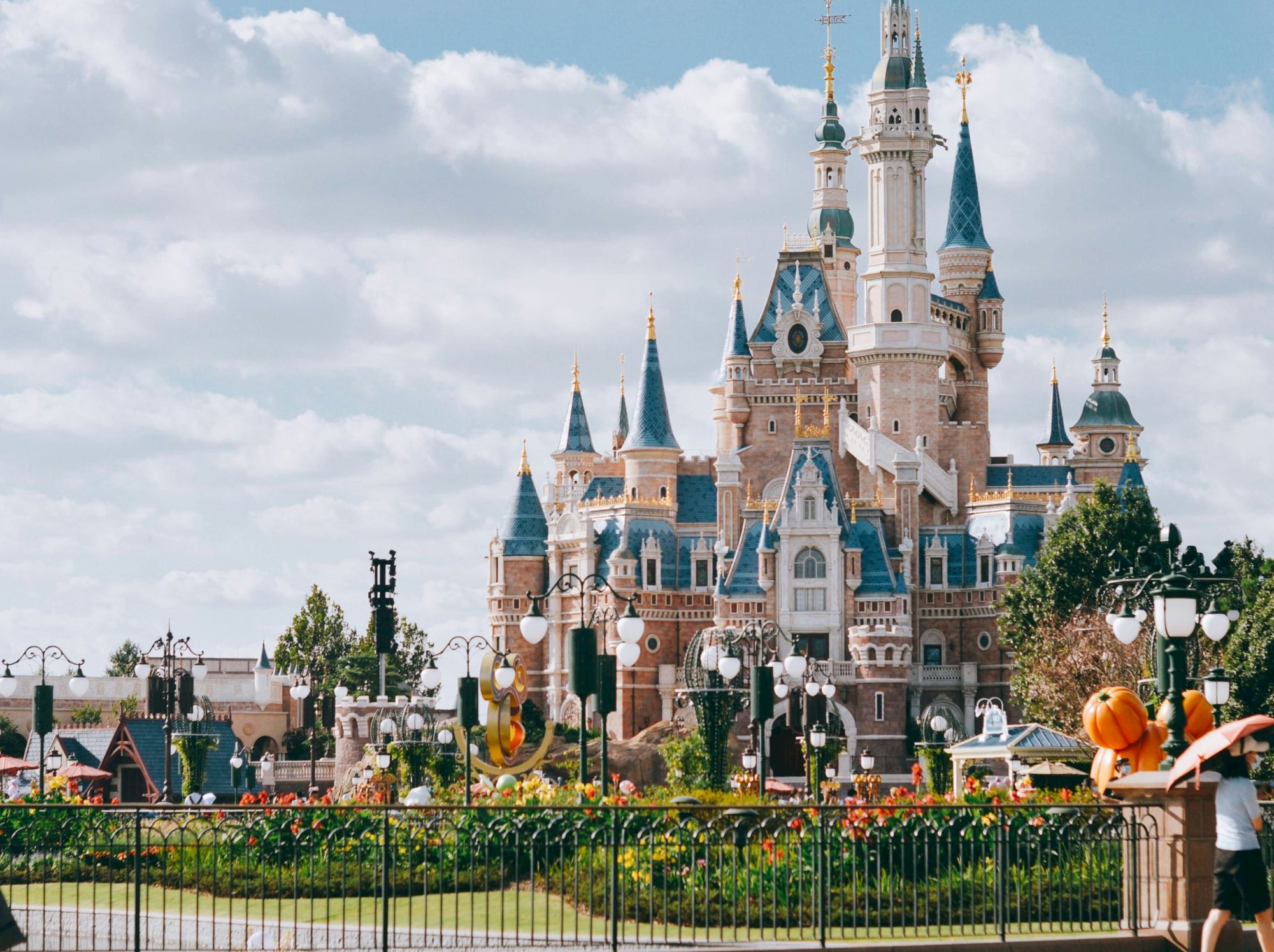 A fairy-tale castle dominates a sunny park with a fountain, trees, and visitors near a wrought-iron fence. Magical, family-friendly vibes.